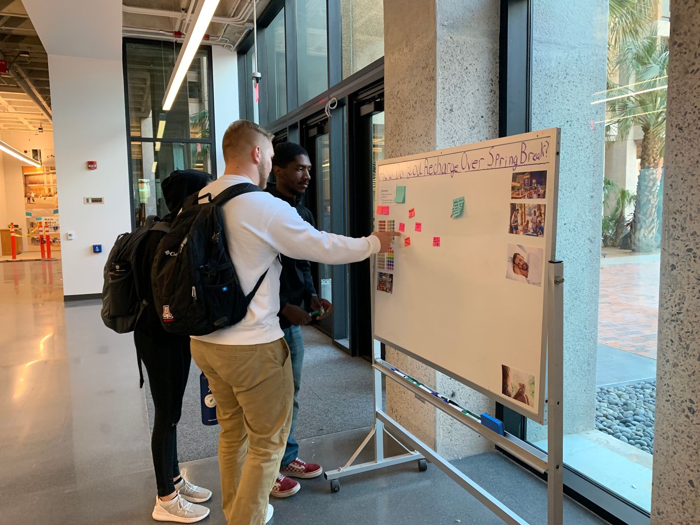 Three students pointing at sticky notes on a whiteboard at the entrance to a public space